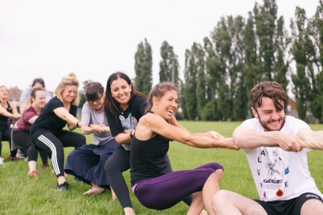 a team of 7 pull a thick rope in a field, they are all smiling