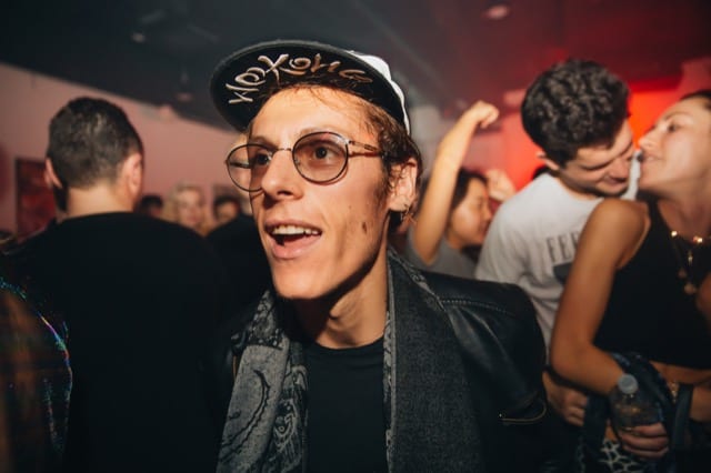 a male with a cap, classes and scarf is smiling away from the camera in a night club