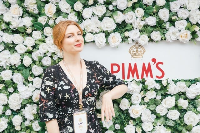 a lady leans on a wall of white roses with Pimms branding on it