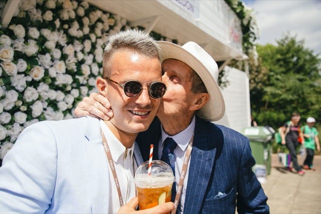 a man kisses another man on the cheek as they enjoy a summer's day in front of a wall of white roses