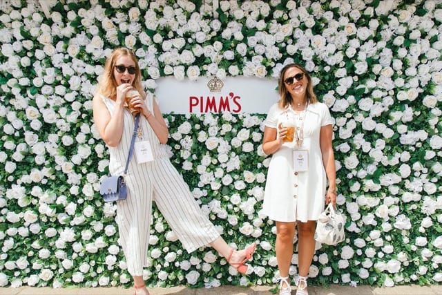 two young ladies smiling each with a cup of Pimms in hand, on a wall of white roses