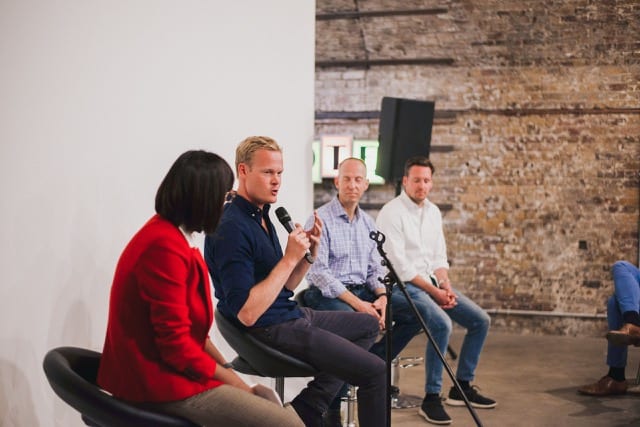 three men and a moderator at a panel, sitting in stools backs to a white wall, one man is answering a question to the audience