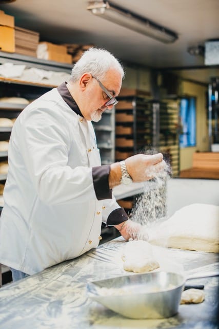 a baker in his work clothes, designer glasses throwing flour on a piece of dough in a modern bakery