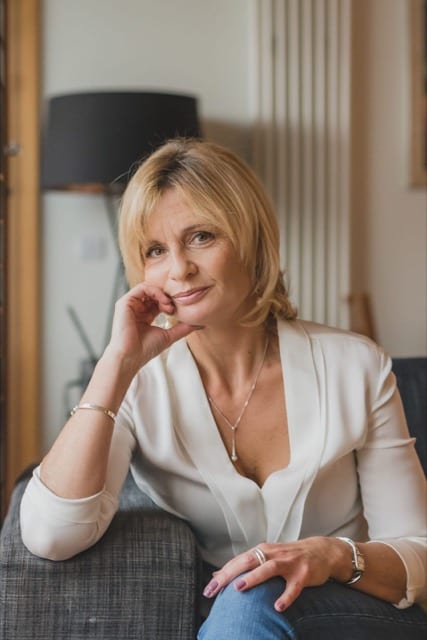 blonde middle aged lady sitting on a sofa with her right on the arm rest, supporting her right cheek