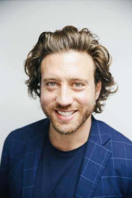 a short bearded man with curly pushed back hair in a blue blazer smiles widely at the camera