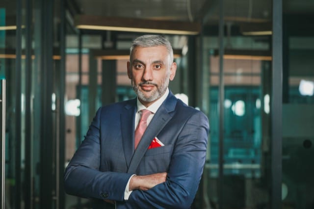 a south asian man with short and tidy white hair and luxurious shiny blue suit smiles at the camera with crossed arms