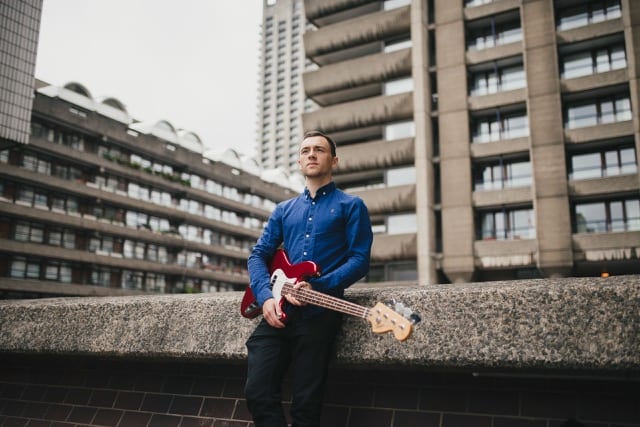 a musician holding an electric guitar leans against a wall in the Barbican