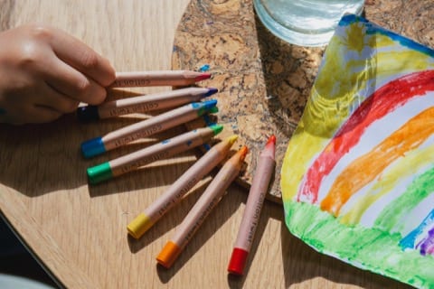 different color crayons laid out in a circle, with a drawing next to it, a hand is reaching for one of them