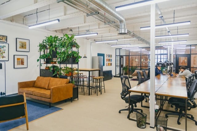 an office displays a row of work tables, a seating area with sofa and racks of plants, black is accented throughout