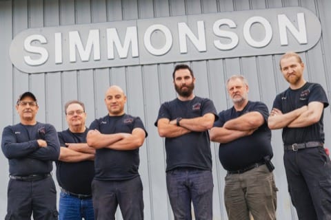 a team of men in work shirts with their hands crossed and smiling towards the camera are infront of the Simmonson building