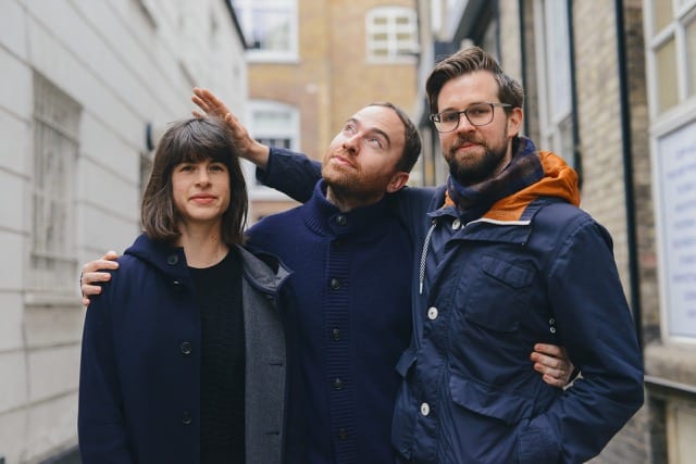 a young team of three with dark hair, outside in a narrow street, each in warm winter navy blue coats, playfully posing