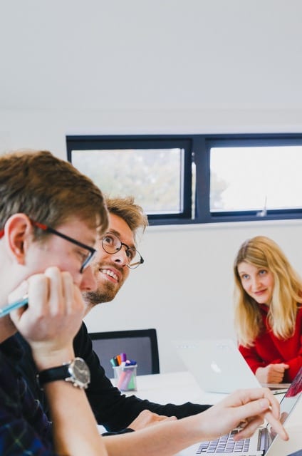 a young team of three within the office, one is looking at another, who is look up at the wall with a bright smile