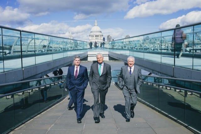 three older whitehaired man in nice suits walk up the ramp of the millenium bridge, st pauls is in the background across the river