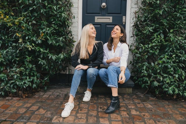 two girls are sitting on a nice doorstep with hedges either side, laughing to the sky