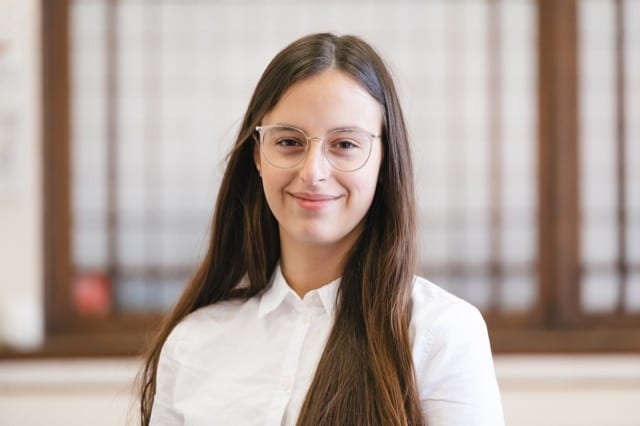 brunette girl with transparent frames smiles at the camera, a brown window frame is blurred in the backgroudn
