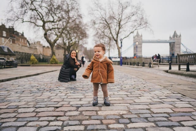 Woman watches her child in a brown coat toddle towards their perfocal photographer near the south bank in London