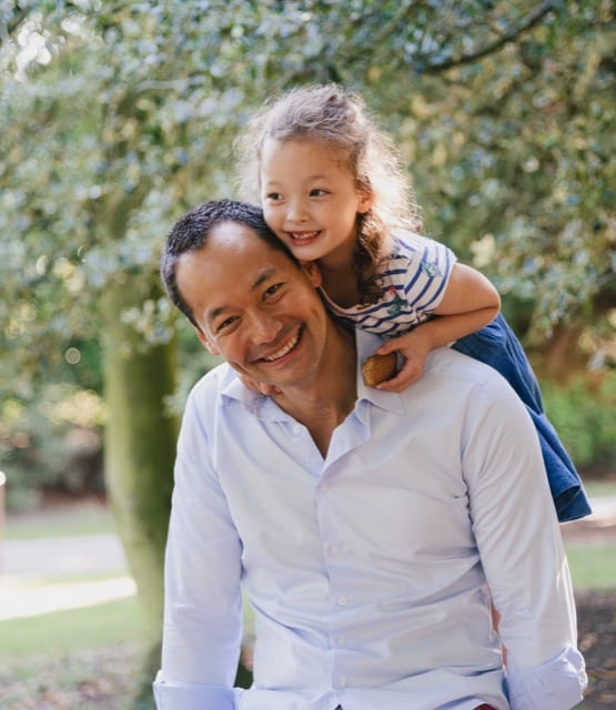 Father and daughter smile for their family photoshoot in Kensington gardens, his daughter climbing onto his shoulders