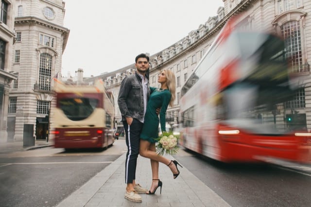 A young couple stand on an island on Regent's st, London for their perfocal couple photoshoot whilst two buses speed past