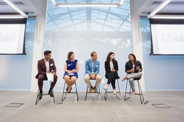 a panel of 5 people sitting on stalls at a business presentation direct a question to the lady at the end of the row