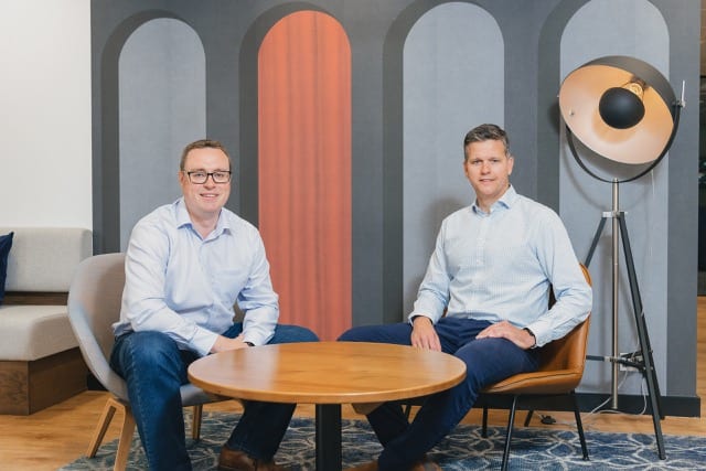 2 men in blue shirts and jeans sitting on small chairs pose for their publicity photoshoot
