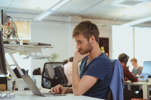 Brown haired man in blue shirt stares thoughtfully at his mac laptop