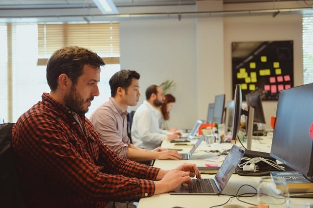 Row of 4 people working at their desks, man in the foreground has a red chequered shirt on