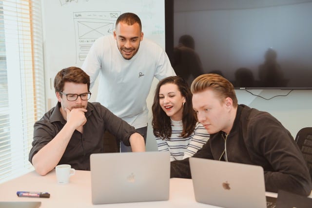Team of 4 people in a meeting room all looking at a man's laptop and laughing
