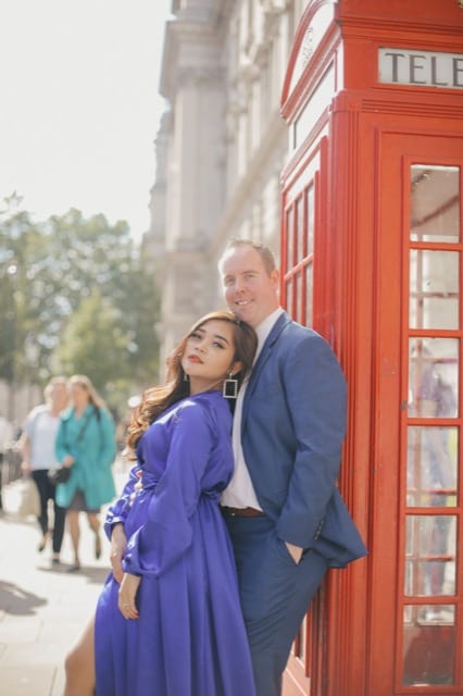 Couple pose for travel photos on London streets in front of red phone box