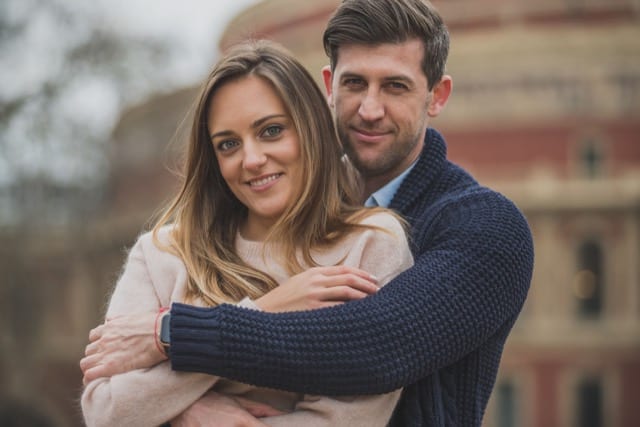 Boyfriend wraps his arm around his girlfriend as they smile into the camera in front of the Royal Albert Hall