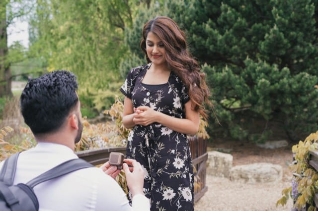 Man on one knee proposing to his girlfriend on a small bridge covered in lilac wisteria