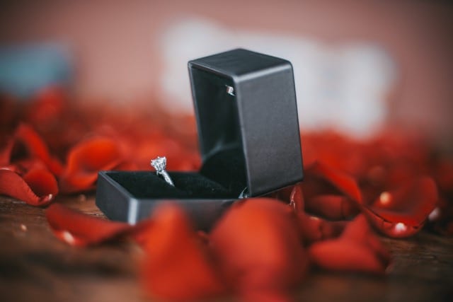 Close up of a diamond engagement ring in its box on a table of red rose petals ready for the perfocal engagement photoshoot