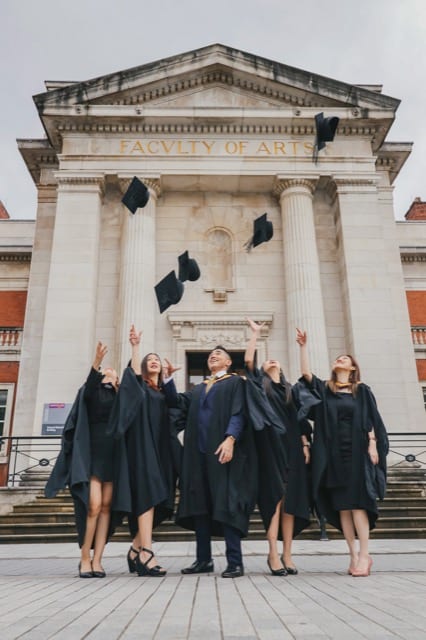 A group of asian graduates throw their caps into the air in front of the faculty of arts building at University of Manchester