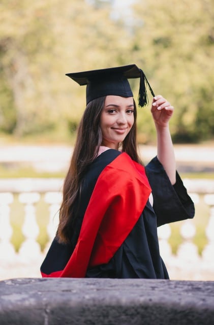 Girl lLook back at the camera and in a graduation gown with red hood and touches tassel of graduation cap
