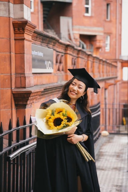 Girl in a graduation cap and gown holds a bunch of sunflowers and smiles at the camera at UCL