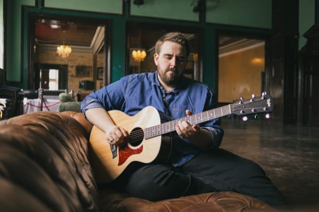 Lifestyle photography of a man in a blue denim shirt playing whis acoustic guitar