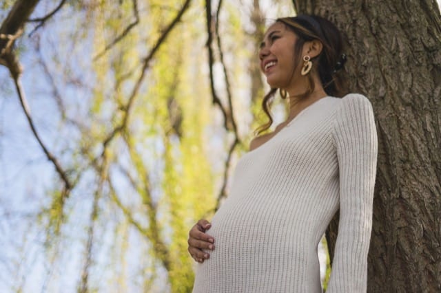 maternity photoshoot in kensington gardens with asian women resting on a tree resting her hand on her pregnant belly
