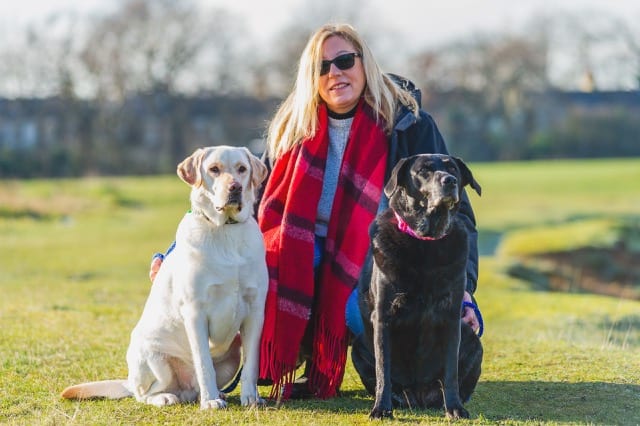 A woman in a red scarf poses in a park with her two labradors; one yellow the other black.