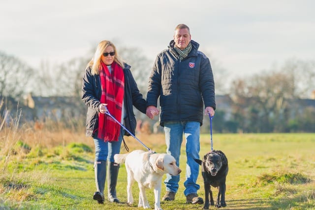 Couple walking their two labrador dogs in a field, one black and the other yellow