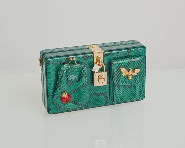 a boxy green reptile skin bag with gold insect brooches on a white glossy table