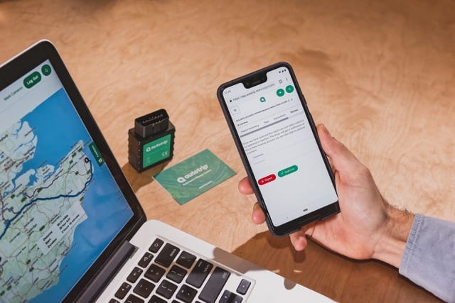 a hand is holding a mobile phone with the autotrip mobile app, a laptop also has the website open in the background
