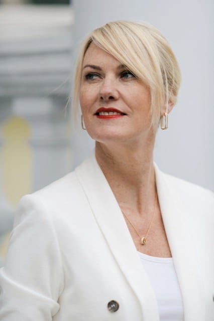 Blonde woman in a white suit looks off and over her shoulder to the distance as she has her headshot taken