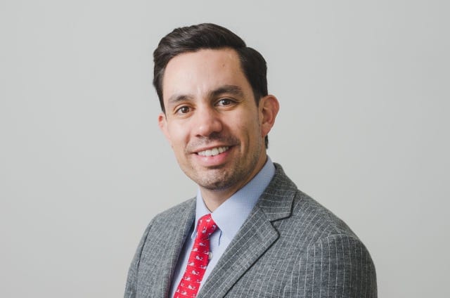 a headshot of a man in a grey pin-striped suit with a blue shirt and red tie