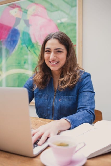 Brunette young woman smiles for her headshot at her computer inside a coffee shop