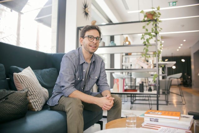 Man in unbuttoned blue shirt sitting on an aquamarine sofa leans forward in conversation for a more candid headshot