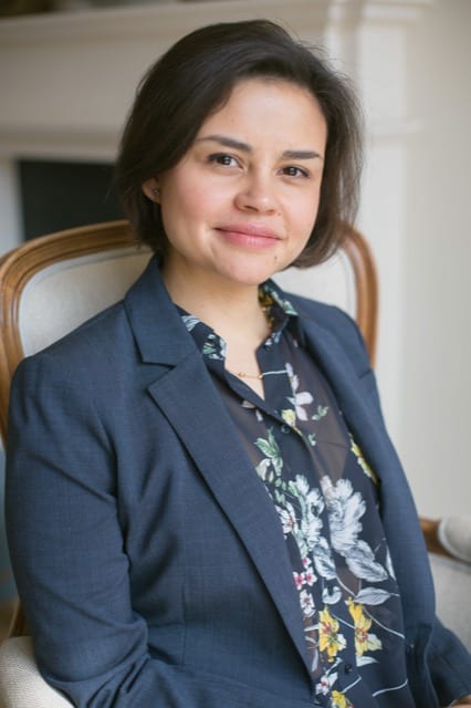 Woman with a brunette bob in a navy suit poses for her headshot by sitting in a grey upholstered chair
