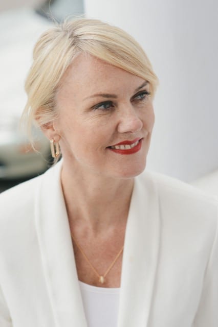 Blonde woman in a white suit looking to her left and smiling for her photo