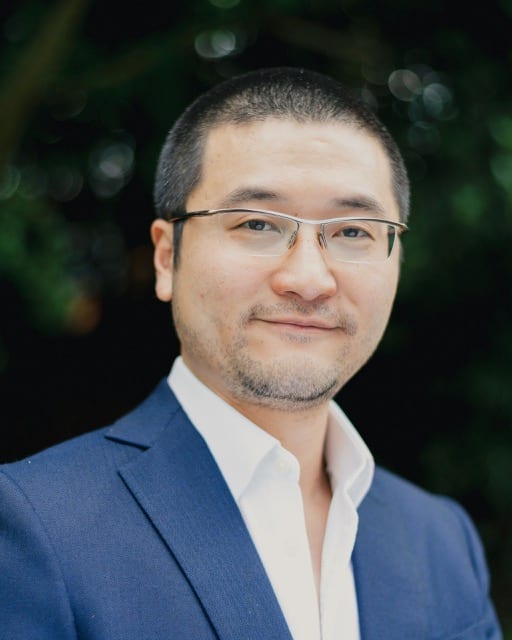 Asian man with light framed glasss poses for his business headshot in a blue suit in front of a dark background