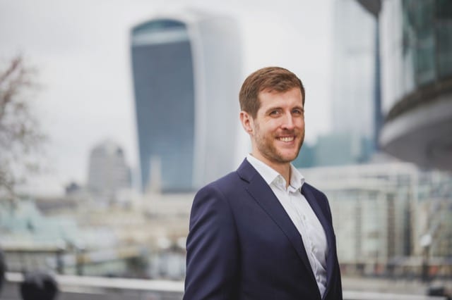man in a suit stands in front of a backdrop of canary wharf