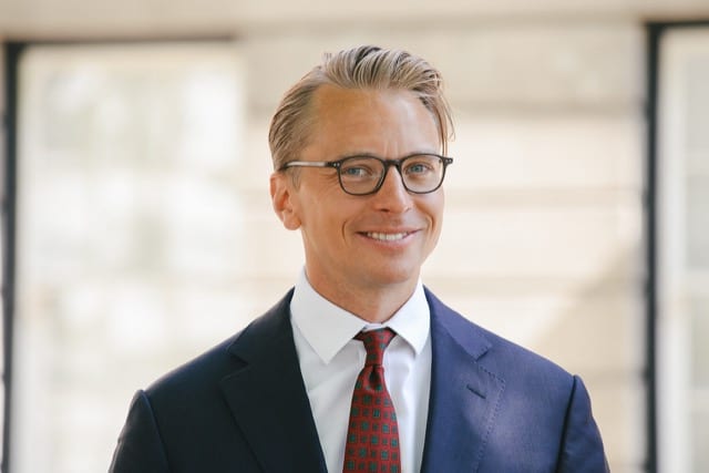 a close up of a blonde man in a suit with a red tie smiling for his headshot