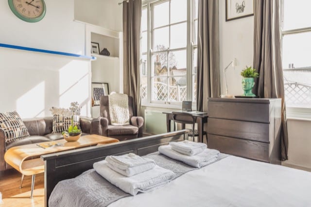 A bedroom with large windows. At the end of a double bed are towels laid out for two as well as a small wooden coffee table and grey sofa